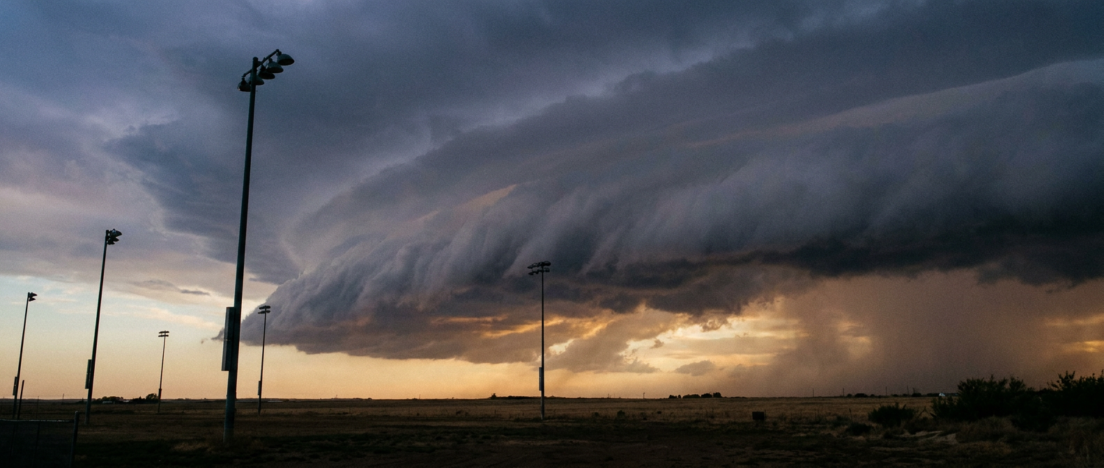 A wide dusk skyscape over a flat grassy outfield-style field with tall stadium light poles in silhouette, a dark shelf cloud advancing from the horizon, warm amber sunset light fading behind it, low camera angle looking up at the approaching cloud line, muted blue-gray and gold color palette