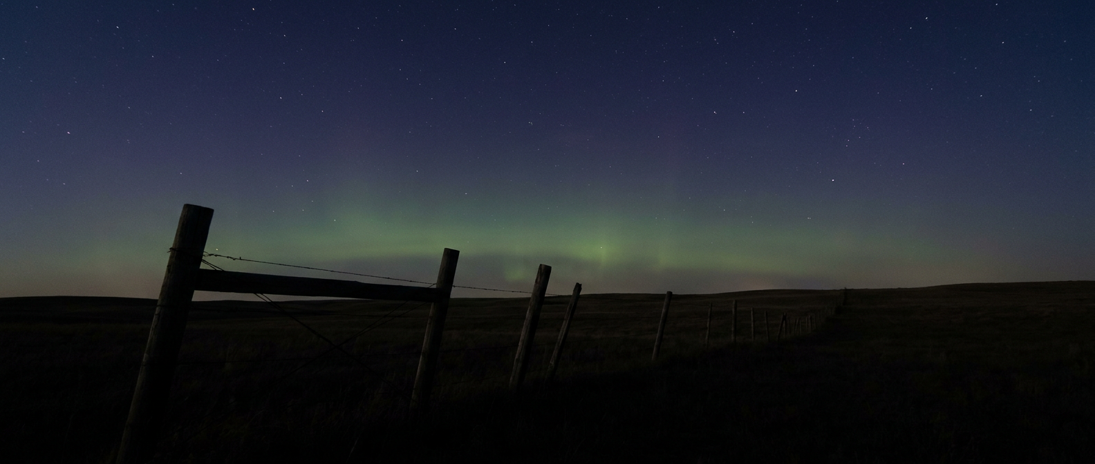 A faint greenish aurora glow low on the northern horizon above a dark rolling prairie, silhouette of a weathered fence line in the foreground, scattered stars overhead, deep indigo and muted green color palette, wide low-angle shot looking north across open grassland