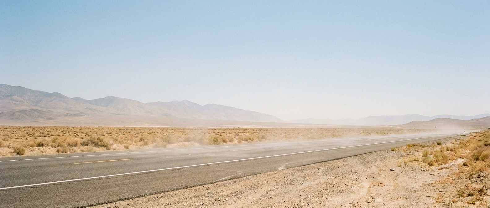 Heat haze shimmering above a lonely two-lane highway crossing high desert scrubland, harsh midday sun overhead, distant mountains fading into a pale dusty haze, warm bleached-out color palette, wide establishing shot from ground level looking down the road