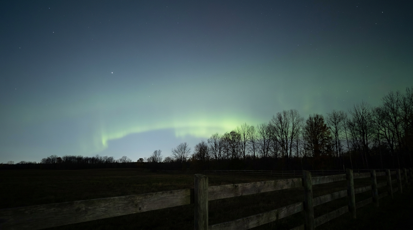 A dark rural horizon at night with a faint band of pale green aurora glow low in the northern sky above a silhouetted line of bare autumn trees, a weathered wooden fence running through the foreground, cool blue-black sky fading to deep green near the horizon, wide-angle shot looking north from ground level