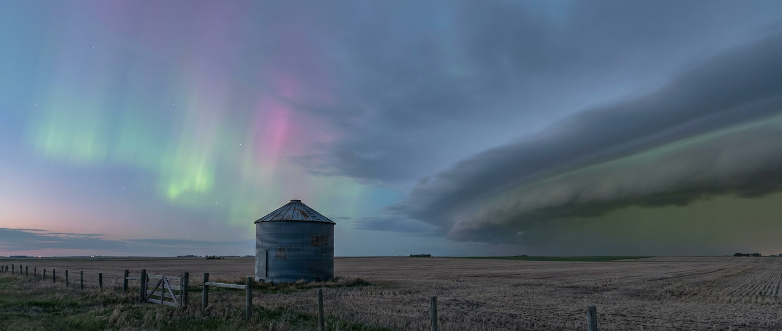 A wide dusk skyscape over open northern prairie farmland, faint green and pink aurora ribbons glowing high in a clear patch of sky on one side, while on the opposite horizon a dark shelf cloud with a greenish undertone advances low over the fields, a lone grain silo and fence line in the foreground for scale, cool blue-gray palette blending into aurora green, wide establishing shot, no text or labels