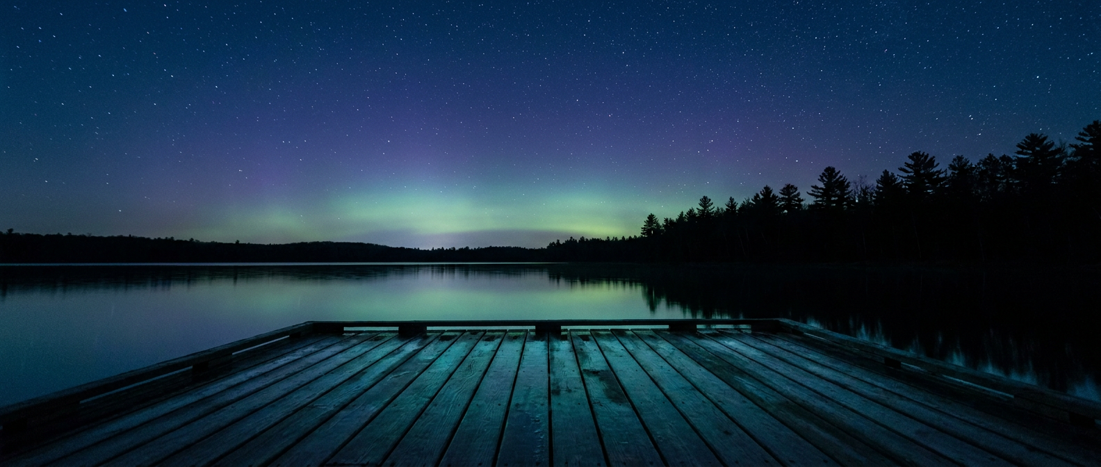 A faint green and violet aurora glow low on the northern horizon above a dark pine forest and a still lake, viewed from a dock, deep blue night sky filled with stars, cool blue-green color palette, wide low-angle shot looking north