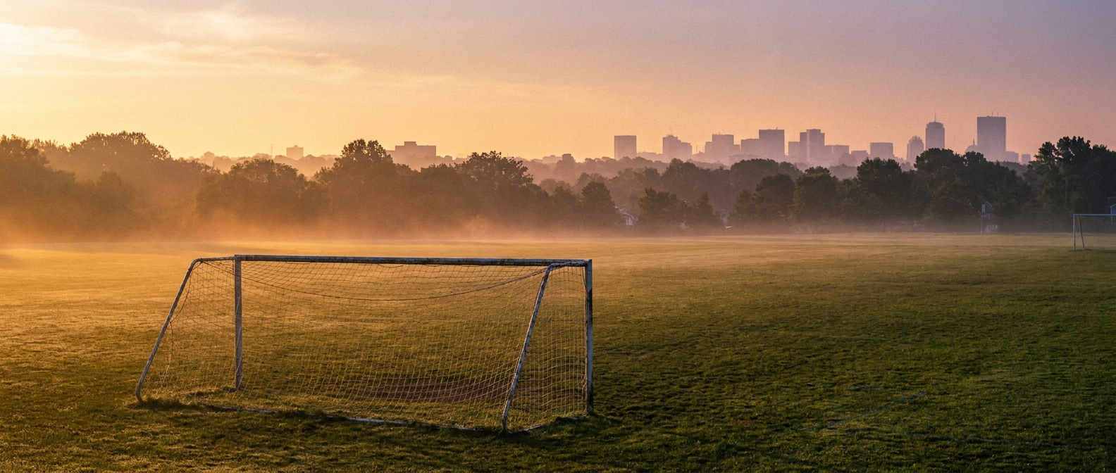 Wide grassy athletic field with a soccer goal in the foreground at golden hour, heat haze shimmering above the turf, a hazy suburban New Jersey skyline silhouette in the distance, warm gold and soft violet color palette, eye-level wide shot