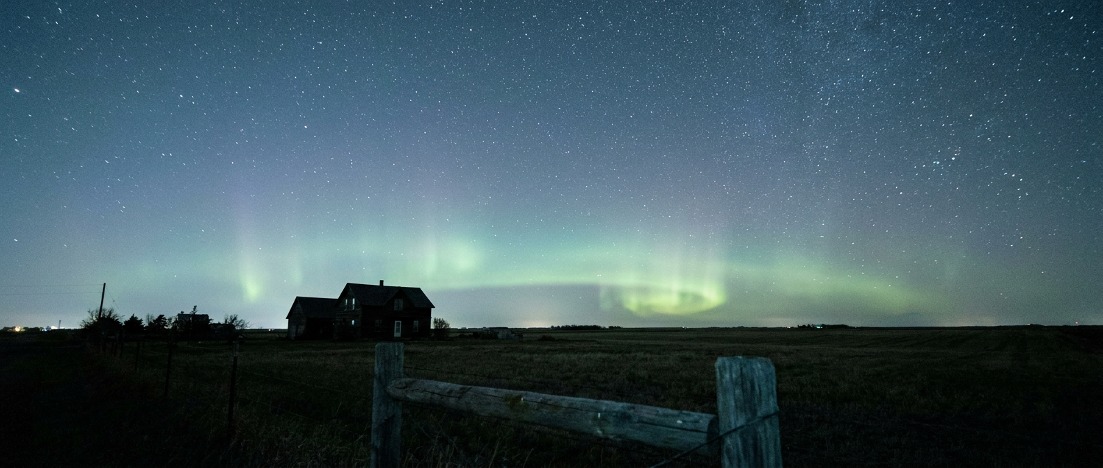 Faint green aurora glow low on the northern horizon above a dark farmhouse silhouette and open field, star-filled sky, cool blue-green palette, wide-angle shot from ground level looking north